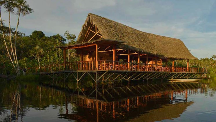 The main building at the Sacha Lodge, with a thatched roof and open-air porch, glows in the afternoon light of the Amazon and reflects in the still water.