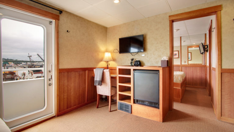 Seating area of commodore suite, wooden desk, with mini fridge, flat screen TV opening to adjoining room aboard the Safari Explorer Hawaii small ship