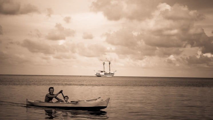 man and son canoeing on still water with the ombak putih small ship in the background with its sails down