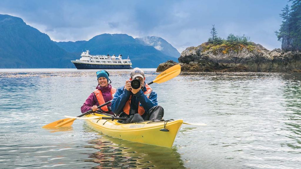 An Alaska small ship cruises while in a double yellow kayak a female paddles while her male partner snaps a photo.