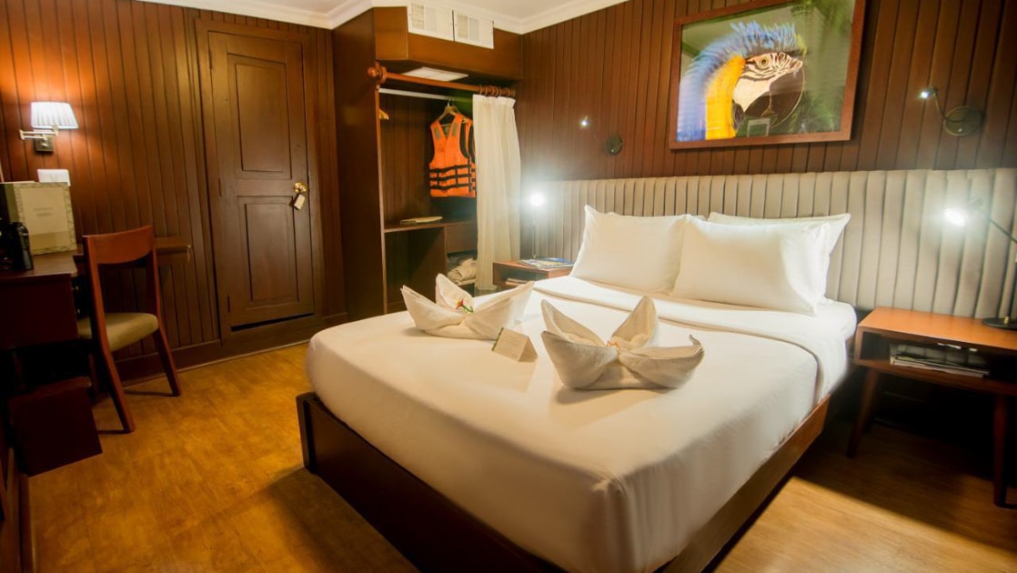 Ship cabin with double bed in white linens, dark wood paneled walls, plush gray headboard, lamps & parrot photo on the Amatista.