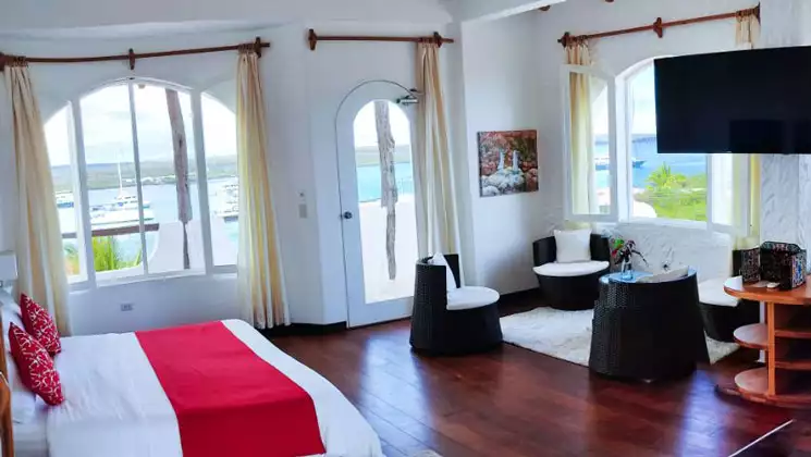 Large bed in white & red linens with 3 chairs beside in white room with wood floors at Angermeyer Waterfront Inn in Galapagos.