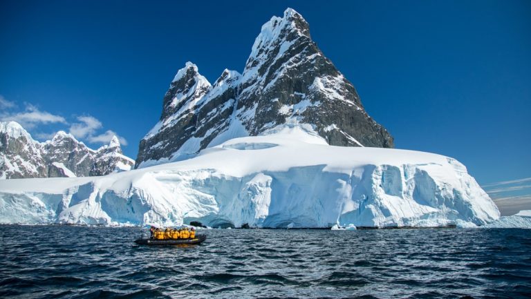 Polar travelers in yellow jackets cruise calm water in a black Zodiac boat beside a snowfield under blue sky in Antarctica.