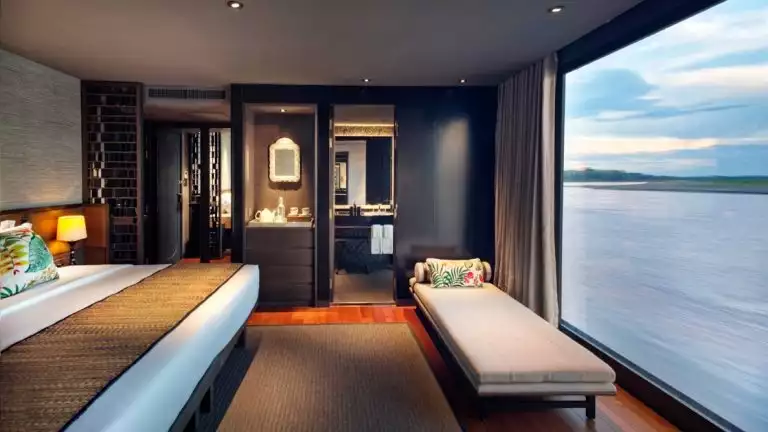 A suite aboard Aqua Nera with a panoramic floor to ceiling window with views of the river outside, king bed and small chaise lounge.