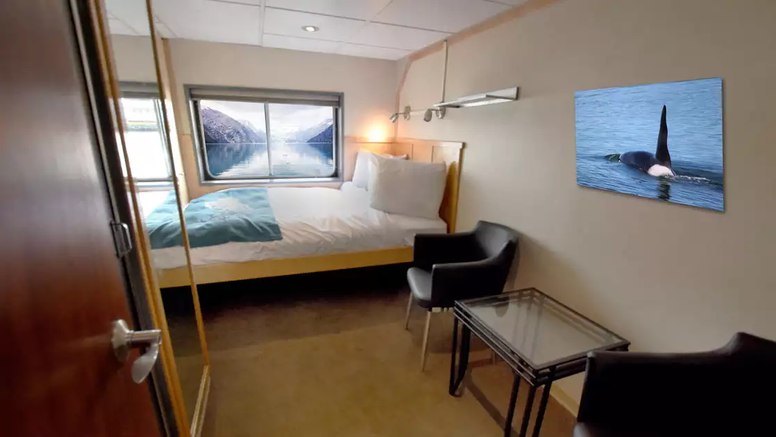 Category A cabin on Baranof Dream Alaska small ship with double bed in white linens, picture window, brown chairs & glass coffee table.