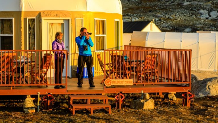 2 Arctic travelers in down jackets stand on wooden deck outside beige yurt at Base Camp Greenland while photographing at sunset.