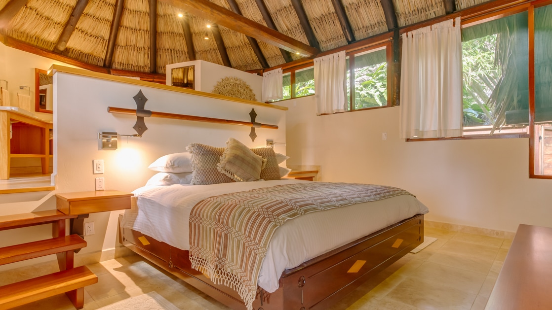 Large bed surrounded by windows looking into the jungle with a thatched roof overhead in the Tree Top Villa at Chaa Creek in Belize.