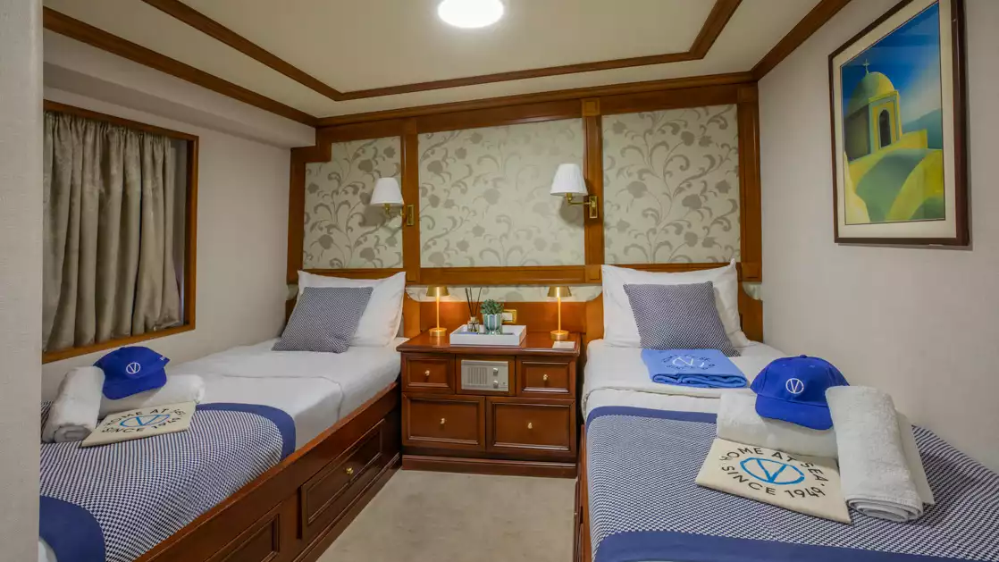 Two twin beds in the Callisto ship category B cabin with yellow pillows and a wicker basket on a bedside table between them