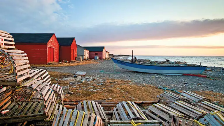 A blue wooden boat, bright red houses and wooden crab pots set along the rocky shoreline of Newfoundland during a pastel sunset.