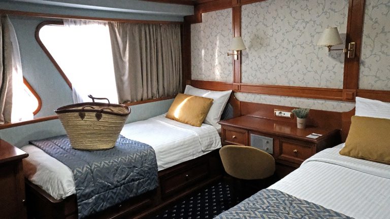 Category A cabin aboard Callisto yacht showing two twin beds with a window and a basket sitting on one bed