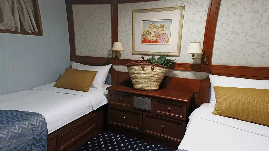 Two twin beds in the Callisto ship category B cabin with yellow pillows and a wicker basket on a bedside table between them