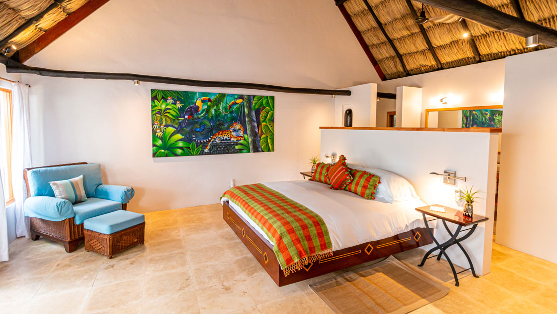 Large bed with white bedding backed by white half-wall in modern room with jungle painting & tile floor at Chaa Creek in Belize.