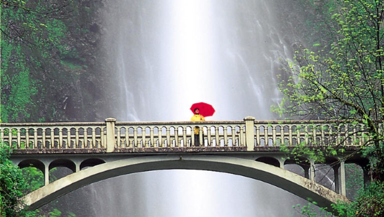a lone traveler with a yellow jacket and red umbrella stands on the multnomah falls bridge with the waterfall in the background