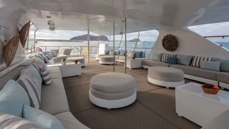 Outdoor top deck lounge on Corals Galapagos ships with padded gray & blue sofas & ottomans & sun cover overhead.