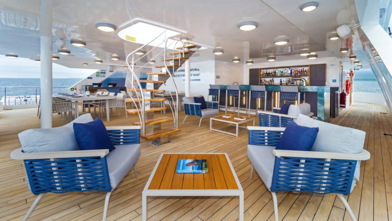 Sundeck outdoor lounge area on Cormorant II ship with padded blue seats, wood coffee table, marble bar, teak decking & stairs.