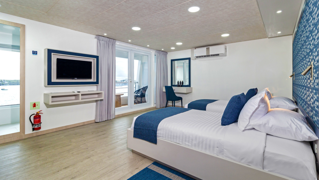 2 twin beds in white & blue face floor-to-ceiling windows on either side of TV & small corner desk on Cormorant II catamaran.