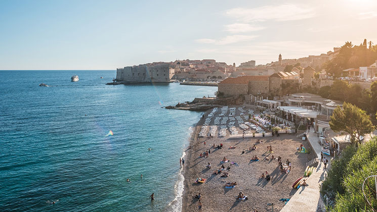 Aerial view of seaside Mediterranean city at sunset, with people on the beach & stone buildings in the background.