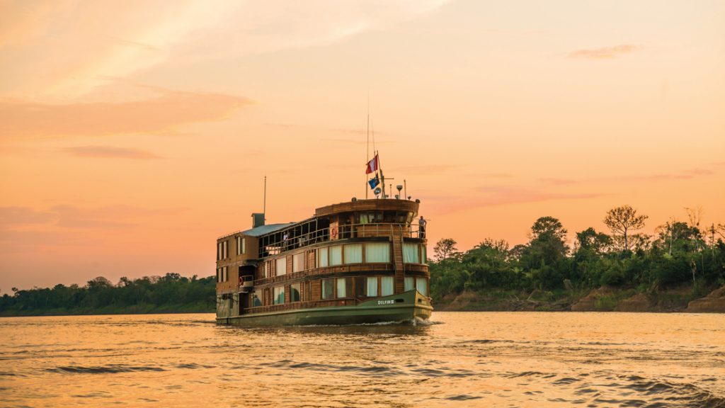 A small river boat cruising down the river with an orange and pink sunset as a backdrop