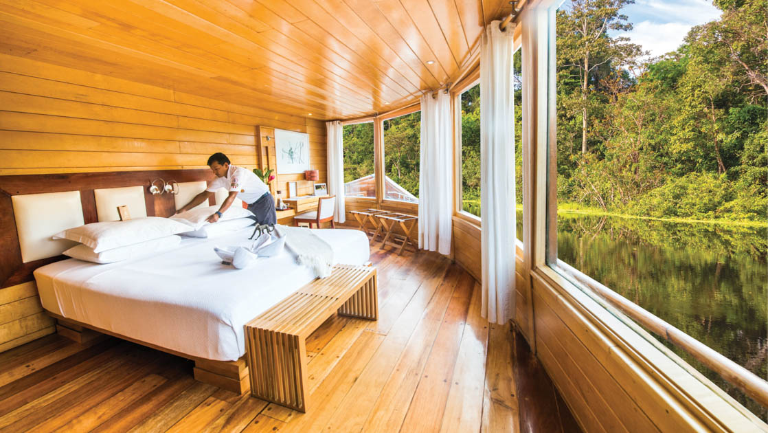Cabin steward makes up king bed by wraparound picture windows in Amazon Suite of Delfin II riverboat on an Amazon River cruise.