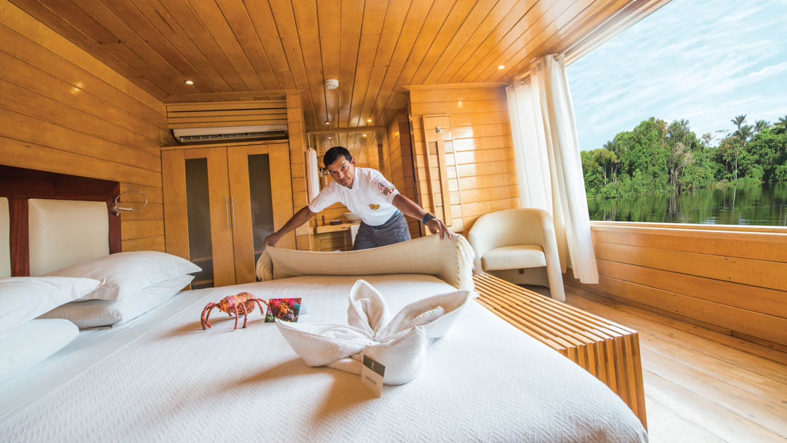 Cabin steward makes up king bed by large picture window in Guest Suite of Delfin II riverboat on an Amazon River cruise.