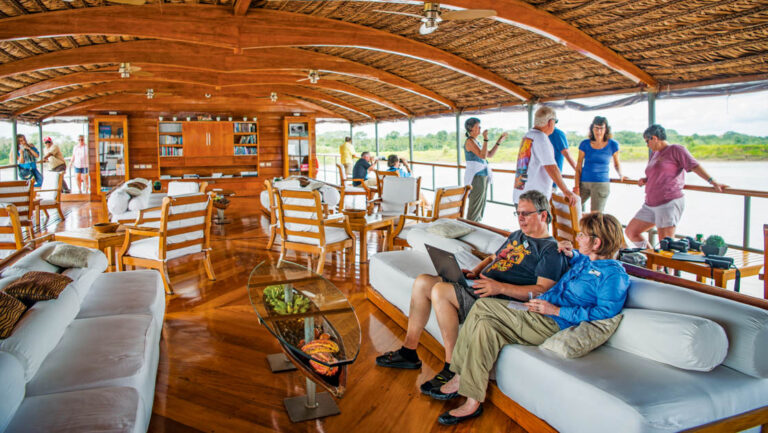 Guests sit on couch & stand along open-air railings in al fresco lounge with couches, chairs & tables on Delfin II riverboat.
