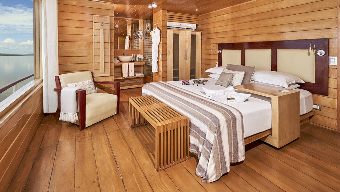 Suite aboard Peru Amazon riverboat Delfin II, showing double bed, chair, ensuite bathroom & wood & white accents throughout.