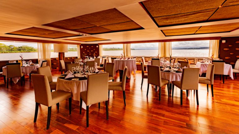 Beautiful wood finishes with white fabric draped tables set for a meal with windows surrounding the room allowing for views of the Amazon