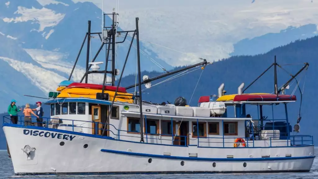 Discovery classic yacht in Alaska with white & blue hull & decks, kayaks aboard & guests on bow looking at mountains.