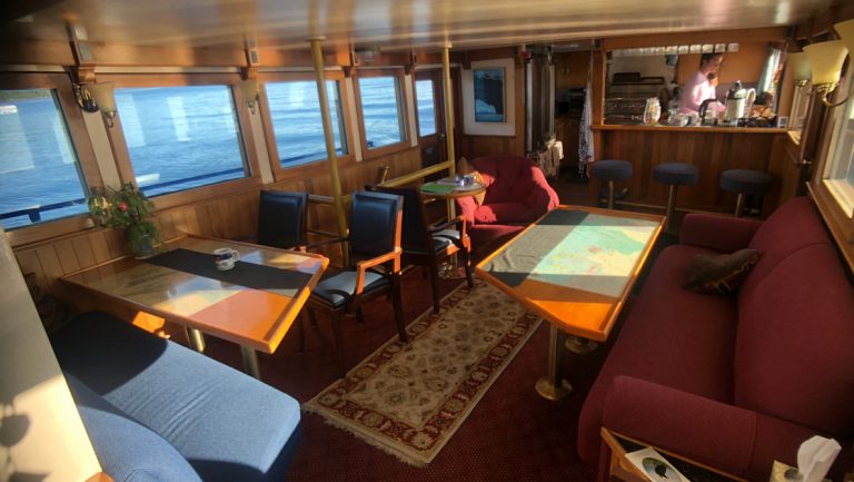 Lounge on Discovery yacht in Prince William Sound, Alaska, with cozy red & blue couches & chairs, wood tables & small bar.