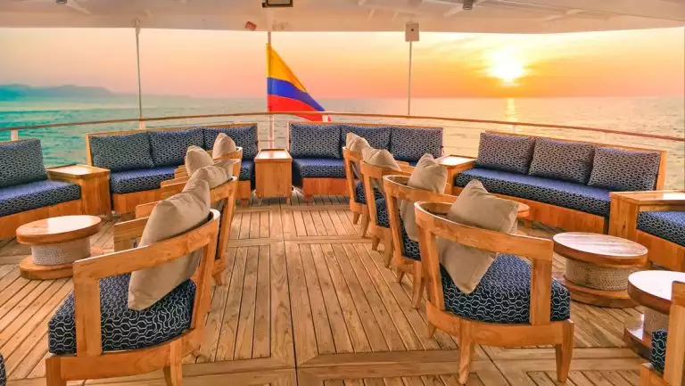 Al fresco lounge on Evolution Galapagos yacht with teak wood decking & wood captain's chairs with blue pillows at sunset.