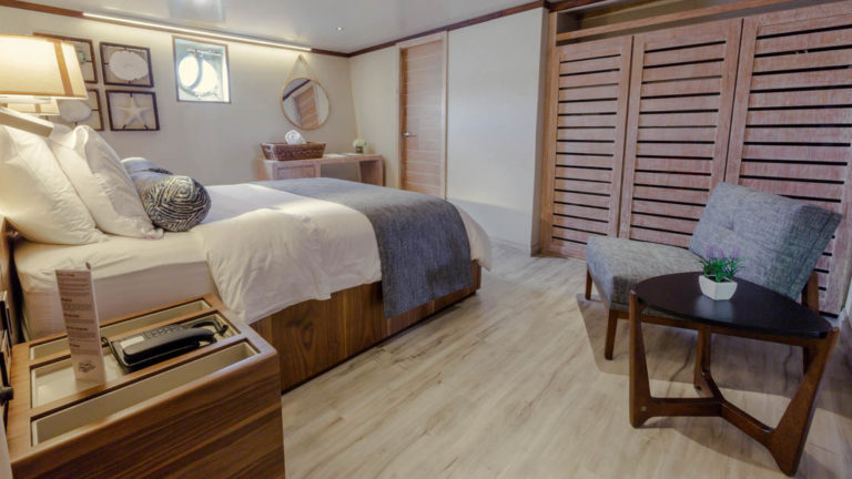 Evolution stateroom with double bed, chair, end table, nightstand and porthole.