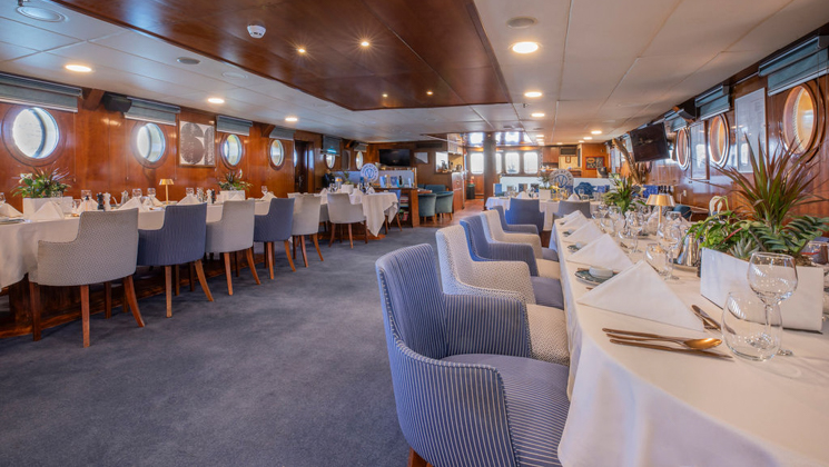 Dining room on Galileo cruise ship with light blue carpet & chairs, tables in white linens, big portholes & green plants.