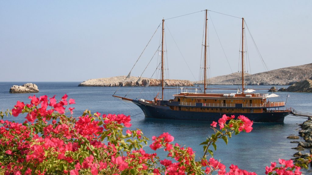 Galileo motorsailer with dark blue hull, teak sun deck & 3 masts sits at anchor in a cove flanked by bright pink flowers.
