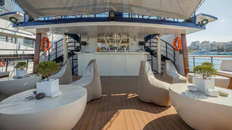 Upper deck outdoor lounge on Galileo ship with solid white tables topped with stones & bonsai trees, plush chairs & white bar.