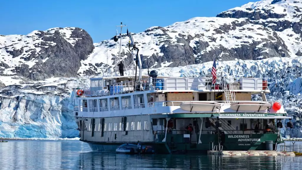 White & green small ship Wilderness Adventure floats close in front of a massive icy glacier on a sunny day.