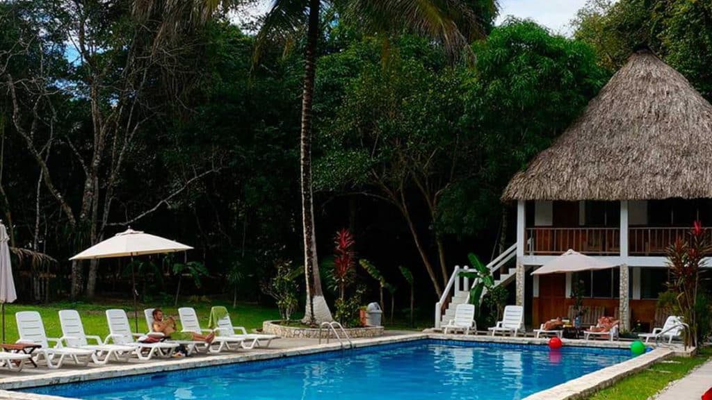 A large pool with lounge chairs and the Tikal Inn a two story building with grass roof.