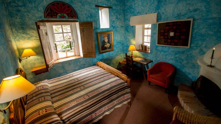 Inside a guestroom at Hacienda Cusin, blue textured walls adorned with Ecuadorian artwork, a fireplace, and a shuttered window to the outsidea stay here as part of an Ecuador land tour,