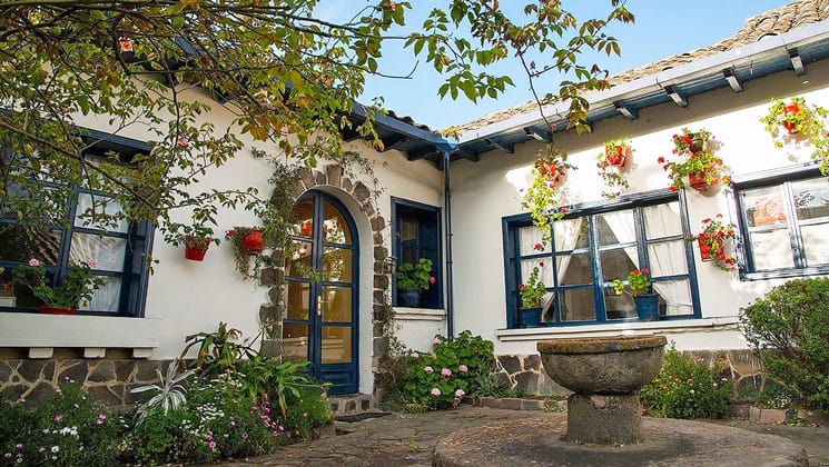 Spotless white walls and blue wooden pained windows of Hacienda Zuleta, seen from a center courtyard with water fountain, from the overhanging roof, brightly colored flowers inside flower pots hang.