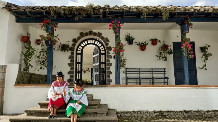 Two Ecuadorian women dressed in green and red traditional clothing woth black hats, sit on the entry steps to Hacienda Zuleta, an estate in Ecuador's highlands. The building has spotless white walls with blue wodden accents and brightly colored potted flowers that hang from the overhanging roof, stay here as part of an Ecuador land tour