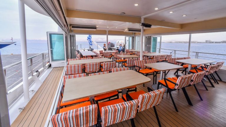 Open-air dining area with empty orange padded chairs with striped backs arranged around various beige tables on Harmony V ship.