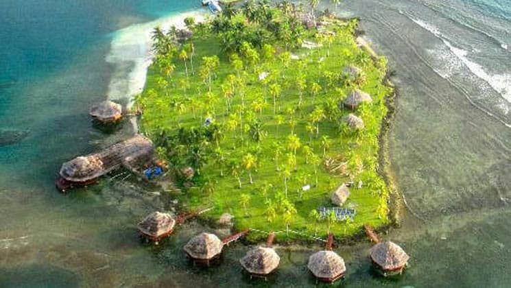 An aerial view of the idyllic eco Hotel Yandup, with thatch-roof cabanas on platforms above the water and adjacent to the island in San Blas, Panama