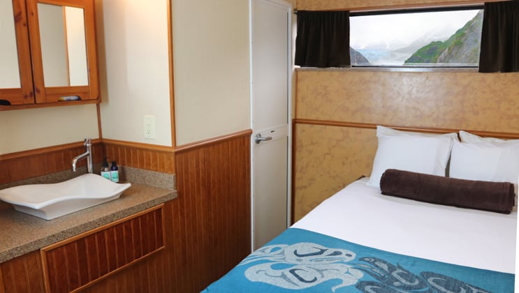 Cabin with queen bed, small window, sink and mirror, with wood accents, on the Kruzof Explorer Alaska small ship.
