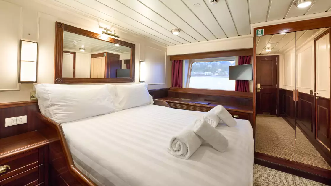 A double bed with white sheets and 2 folded towels on a small cruise ship with brown interior.