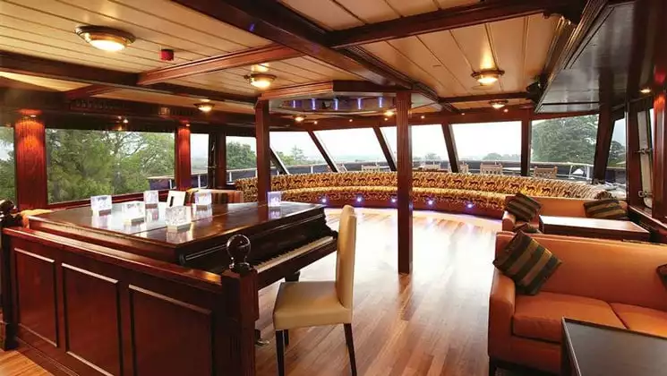 Lord of the Glens ship lounge area with piano, cushioned seating, tables and large picture windows throughout.