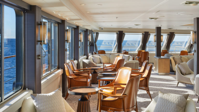 lounge area with tan leather accent chairs, grey upholstered love seats and wide wrap around windows aboard Magellan Explorer luxury Antarctica expedition ship