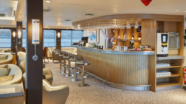 Bar & lounge on Magellan Explorer, ship with wooden bar, stools & wine glasses hanging overhead.