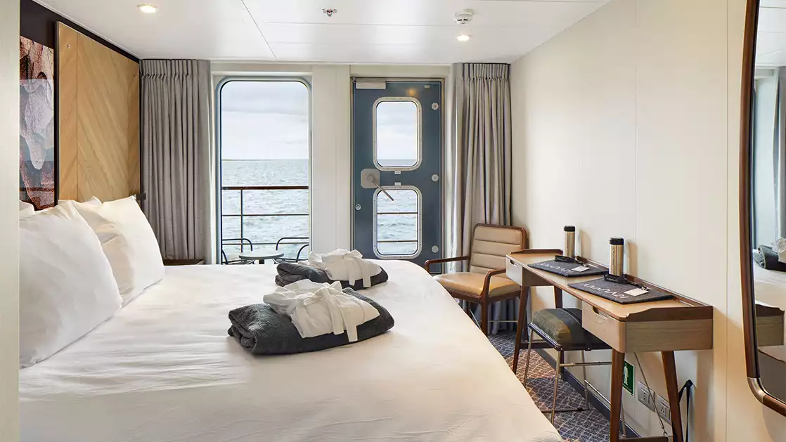 Single Veranda Cabin aboard Magellan Explorer with king bed across from desk with cushioned stool & chair in corner overlooking balcony