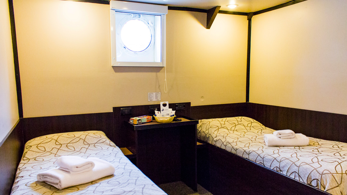 two twin beds with a night stand between them with individual plugs and a welcome basket featuring a porthole above aboard Ushuaia