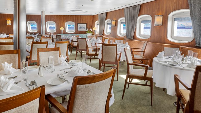 A beautiful dining room featuring handsome wood trim and white linen service aboard the National Geographic Explorer awaits