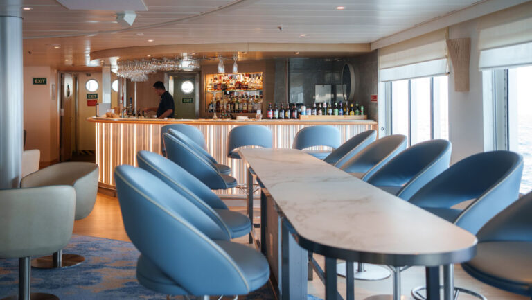 Long table with tall blue swivel chairs leads to modern, lit-up bar by wall of windows on National Geographic Explorer ship.